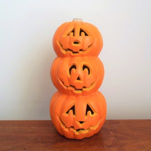May include: A stack of three orange ceramic pumpkins with carved jack-o'-lantern faces. The pumpkins are stacked on top of each other, with the largest pumpkin on the bottom and the smallest pumpkin on top.