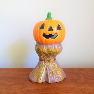 May include: A ceramic Halloween pumpkin with a jack-o'-lantern face sits atop a brown and gold straw base.
