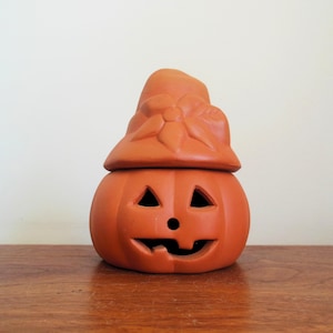 May include: A terracotta-colored jack-o'-lantern with a hat-shaped lid. The pumpkin has triangular eyes, a round nose, and a carved mouth. The hat features a bow detail. This decorative piece is perfect for Halloween.