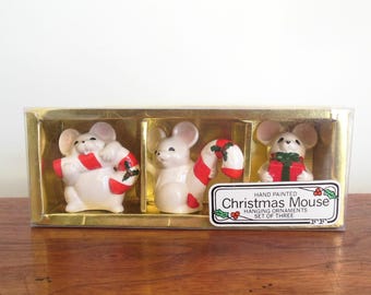 May include: Set of three hand-painted ceramic Christmas mouse ornaments. Each mouse is holding a candy cane or a gift box. The ornaments are packaged in a clear box with a gold background and a label that reads "Hand Painted Christmas Mouse Hanging Ornaments Set of Three".