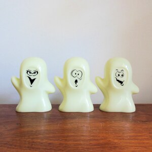 May include: Three yellow plastic ghost figurines with black facial expressions. The ghosts are standing on a wooden surface.
