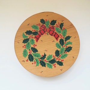 May include: A round wooden box with a painted Christmas wreath design. The wreath is green with red berries and a red and black bow.