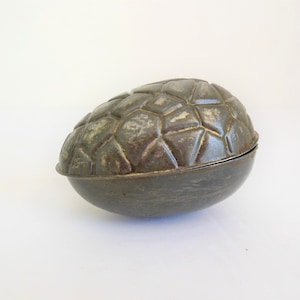 May include: A vintage, egg-shaped metal container with a textured, geometric pattern on the top half. The container is a dark, aged gray color, suggesting it is antique. The bottom half is smooth and rounded.