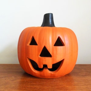May include: Orange ceramic jack-o'-lantern with triangular eyes and a carved, smiling mouth. The pumpkin has a black stem and is a classic Halloween decoration.
