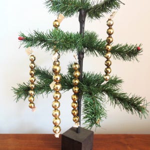 May include: A small, artificial Christmas tree with green needles and gold bead ornaments. The ornaments are strung together and hang from the branches. The tree is set on a dark brown square base. A festive holiday decoration.