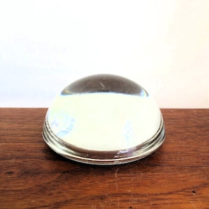 May include: A clear glass magnifying dome with a silver rim. The dome is sitting on a wooden surface.