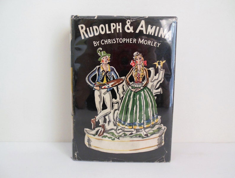 May include: A black book cover with a colorful illustration of a man and woman in traditional clothing. The title of the book is "Rudolph & Amina" by Christopher Morley.