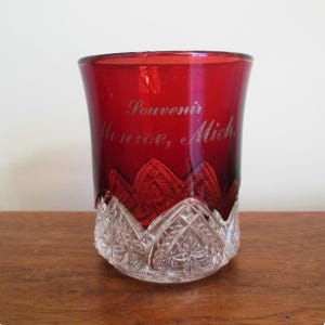 May include: A red glass souvenir cup with a clear, cut-glass base. The cup has the words "Souvenir, Muskegon, Mich." etched on the side.