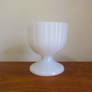 May include: White milk glass vase with a fluted design. The vase has a wide, round base and a tall, narrow body.