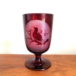 May include: A ruby red glass goblet with a frosted circle design featuring a silhouette of a bird perched on a branch.