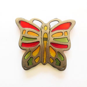 May include: A butterfly-shaped ornament featuring a metallic frame with cutouts filled with translucent red, yellow, and green panels. The metal has a muted, weathered finish, and the design includes symmetrical wing patterns and a central body.