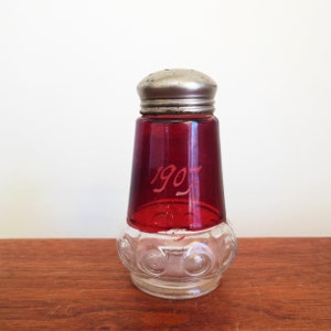 May include: A vintage red glass salt shaker with a silver top and a clear glass base. The shaker has a date of "1907" etched on the side.