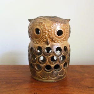 May include: A ceramic owl figurine in shades of brown and tan. The owl has large, round eye holes and a series of circular cutouts on its body. The owl is sitting on a wooden surface.