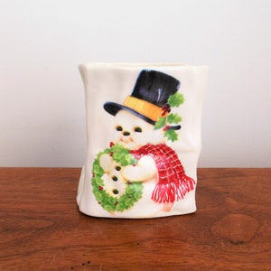 May include: White ceramic Christmas-themed vase with a snowman holding a wreath. The snowman is wearing a black top hat with a yellow band, a red and white plaid scarf, and a green holly sprig.
