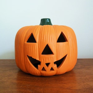 May include: Orange ceramic jack-o'-lantern with a carved smiling face. The pumpkin has a green stem and is a traditional Halloween decoration.