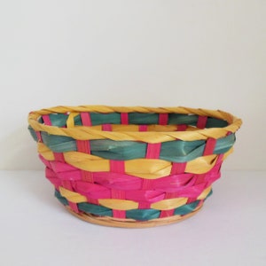 May include: A woven basket with a round shape, featuring a vibrant color scheme. The basket is made of interwoven strips of pink, teal, and yellow material. The rim is reinforced with a braided yellow and teal trim. The base is a natural, light brown color.