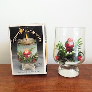 May include: A clear glass candle holder with a Santa Claus figurine surrounded by greenery and red berries. The box next to it reads "Flam'buoyants" and "Santa Claus Candle". The candle holder has a white base.