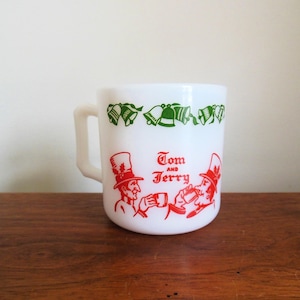 May include: White ceramic mug with a handle, featuring green bell illustrations and red text that reads "Tom and Jerry." The mug has two cartoon figures in top hats. A vintage-style collectible.