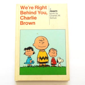 May include: A vintage book titled "We're Right Behind You, Charlie Brown" by Charles M. Schulz. The cover features Charlie Brown, Lucy, Linus, and Snoopy. The characters are standing on green grass against a light blue background.
