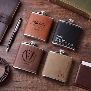 May include: Four personalized flasks with stainless steel tops and leather-like exteriors. The flasks are brown, black, and tan, with engraved text and designs. One flask says "Nicholas Best man 30.10.2025". Another says "David Williams Best Man 22.09.25".