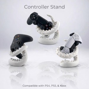 Könnte beinhalten: Drei weiße Controller-Ständer in Form von Krakenarmen, die jeweils einen Gaming-Controller halten. Der Text Controller Stand steht oben und Compatible with PS4, PS5, & Xbox unten. Die Ständer verfügen über detaillierte Saugnäpfe.