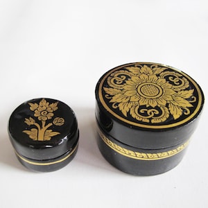 May include: Two black round boxes with gold floral designs. The smaller box has a single flower design, while the larger box has a more intricate design with a larger flower in the center.