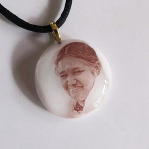 May include: A round pendant with a sepia-toned portrait of a person, suspended from a black cord. The pendant has a gold-colored bail. The portrait is a detailed image of a person's face.