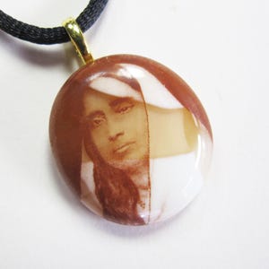 May include: A brown and white oval pendant with a picture of a woman wearing a white head covering. The pendant is on a black cord.