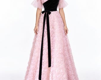 May include: A floor-length gown featuring a light pink textured floral skirt and bodice with off-the-shoulder sheer tulle sleeves. A contrasting black velvet diagonal panel covers the upper bodice, accented by a long black velvet ribbon sash at the waist.
