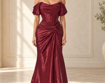 May include: A floor-length, off-the-shoulder gown in a deep burgundy satin fabric. The bodice features a structured corset-style design with gathered fabric, while the skirt includes a central knot detail that creates cascading folds and a train.