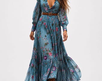 May include: A long-sleeved, floor-length blue dress featuring a floral pattern in shades of pink, purple, and green. The dress has a deep V-neckline with ruffled trim, long sleeves with cuffs, a gathered waist with a brown belt, and a tiered skirt.