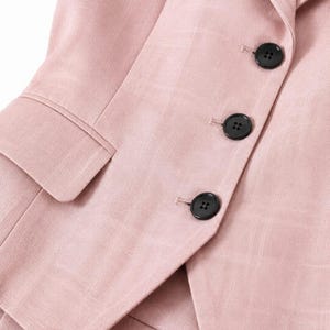 May include: A light pink blazer featuring a subtle grid pattern, a notched lapel, and three black buttons along the front closure. The garment includes a flap pocket on the side and is constructed with a visible textured fabric weave.