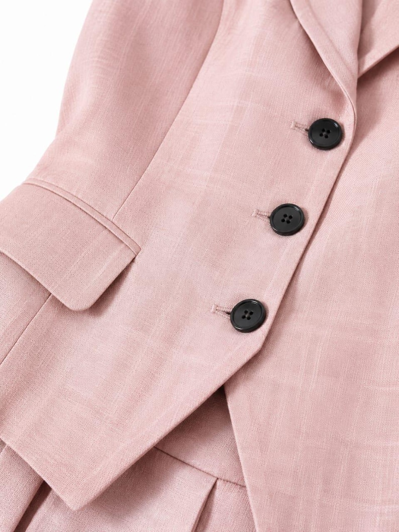May include: A light pink blazer featuring a subtle grid pattern, a notched lapel, and three black buttons along the front closure. The garment includes a flap pocket on the side and is constructed with a visible textured fabric weave.