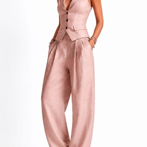 May include: A dusty rose sleeveless vest with a notched lapel and three black buttons, paired with matching high-waisted wide-leg trousers featuring front pleats and side pockets. The ensemble is crafted from a textured fabric with a subtle sheen.