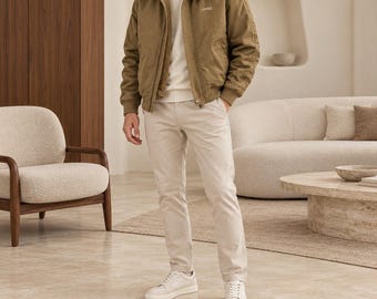 May include: A tan zip-up jacket with a high collar and the word Jeep embroidered on the chest, worn over a white long-sleeve shirt and light-colored trousers. The jacket features a textured lining visible at the collar and long sleeves.