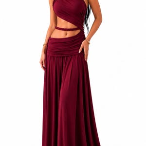 Puede incluir: Un vestido largo de color burdeos con diseño de un solo hombro con tirante trenzado, una abertura en la cintura y una banda trenzada horizontal. La prenda cuenta con un corpiño fruncido y una falda larga y fluida con pliegues suaves.