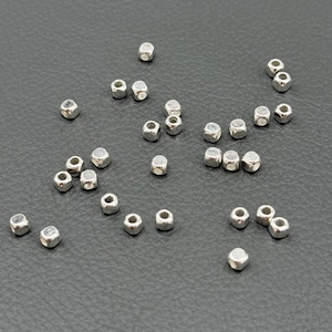 Puede incluir: Cuentas hexagonales plateadas dispersas. Las cuentas son pequeñas, con un brillo metálico, y parecen ser de metal. Están dispersas sobre una superficie gris oscuro, lo que sugiere que son componentes sueltos para la fabricación de joyas o manualidades.