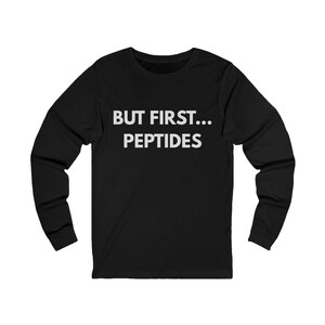 Puede incluir: Camiseta negra de manga larga con el texto blanco "BUT FIRST... PEPTIDES". El texto está en negrita y sin serifa. La camiseta tiene cuello redondo y mangas largas.