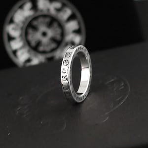 Può includere: Un anello d'argento con scritte incise attorno alla fascia. L'anello è esposto su una superficie scura, con un elemento decorativo sfocato sullo sfondo. Il design dell'anello è semplice ed elegante.