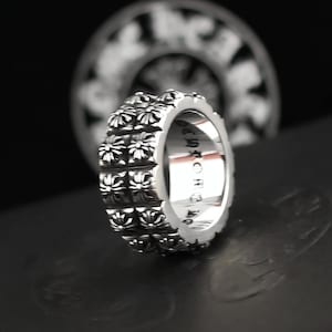 May include: A silver ring with a repeating floral pattern. The ring has a thick band with a series of embossed flowers. The inside of the ring has engraved text. The background is dark, with a blurred image of a similar design.