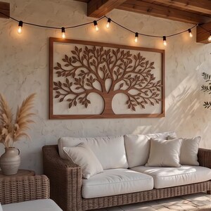 May include: A wooden tree wall art with a rectangular frame hangs above a white sofa with beige pillows. The tree design features intricate branches and leaves. A string of lights hangs above the art, illuminating the outdoor space.