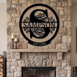 May include: Black metal family name sign with the name "SAMPSON" and the year "EST 1998" cut out. The sign is circular with a decorative letter "S" in the centre. It is mounted on a stone fireplace.
