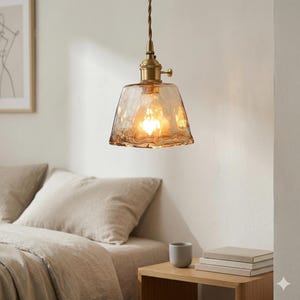 May include: A hanging pendant light with a textured, amber glass shade and brass-coloured metal fittings. The light hangs from a brown cord. The illuminated light casts a warm glow. The light is above a bed and a nightstand. The room is decorated in neutral tones.