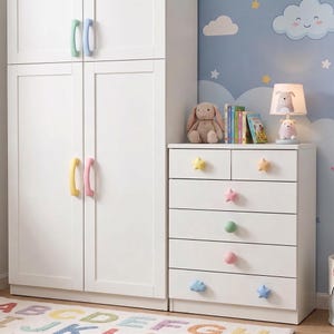 May include: White bedroom furniture set with colorful accents. A tall wardrobe with pastel handles and a dresser with star and cloud-shaped knobs. A bunny lamp and stuffed animal sit on top of the dresser.