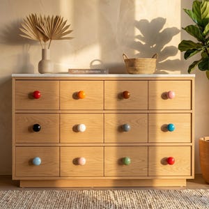 May include: A light wood dresser with twelve drawers, each with a different colored knob. The knobs are red, orange, brown, pink, black, white, gray, blue, light blue, pink, green, and red. The dresser has a white top.