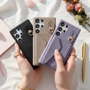 Puede incluir: Tres fundas de teléfono de cuero en negro, beige y lila. Cada funda presenta un acabado texturizado, un recorte para la cámara y un soporte de anillo dorado. Las fundas se muestran sobre una superficie blanca con un cuaderno, flores y un bolígrafo.