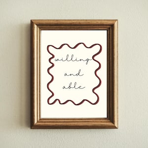 May include: A framed print with the words "willing and able" in black script. The text is centered on a cream-colored background, framed by a wavy, dark red border. The frame is a warm, medium brown color.