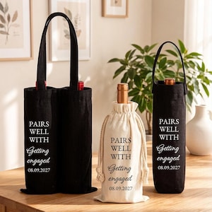 Op de afbeelding: Een zwarte dubbele wijntas en twee enkele wijntassen, één zwart en één crème, allemaal met de tekst "PAIRS WELL WITH Getting engaged 08.09.2027". De crèmekleurige tas heeft een trekkoordsluiting met dikke touwbanden.