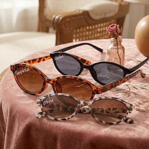 May include: Three pairs of sunglasses with different frame styles and colors are displayed on a pink surface. One pair has a tortoise shell pattern, another is black, and the third is a gray and white animal print. The names "Sophia", "Chloe", and "Olivia" are written on the lenses.