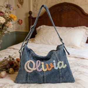 May include: A denim shoulder bag with a long strap. The word "Olivia" is embroidered on the front in pastel colors. The bag is on a bed with a floral pattern.